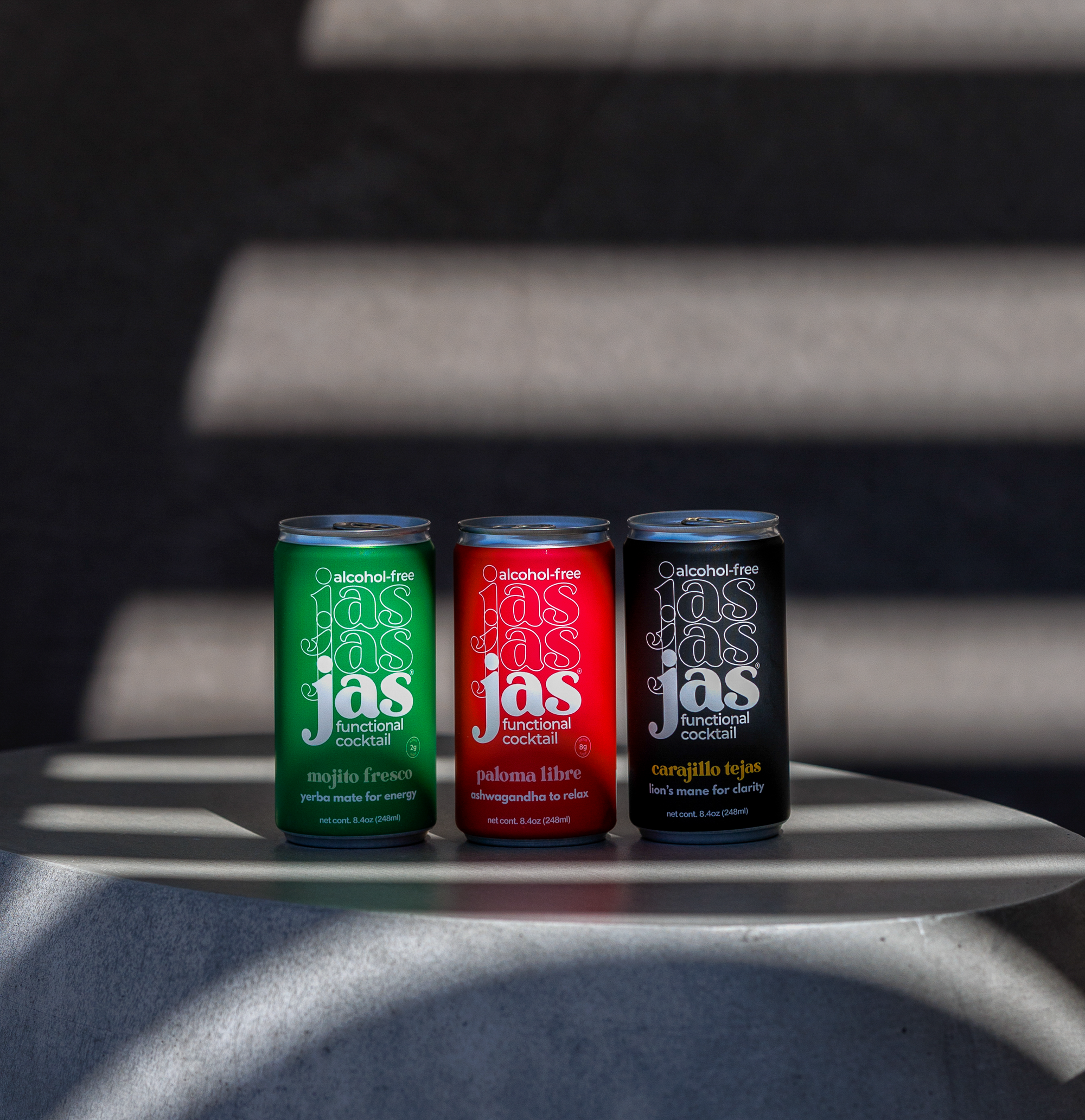 Three slim cans of Jas Non-alcoholic Cocktails sit on a round metallic table under dramatic diagonal shadows. The cans are green, red, and black, each with the brand name 'jas' in a stylized white script.