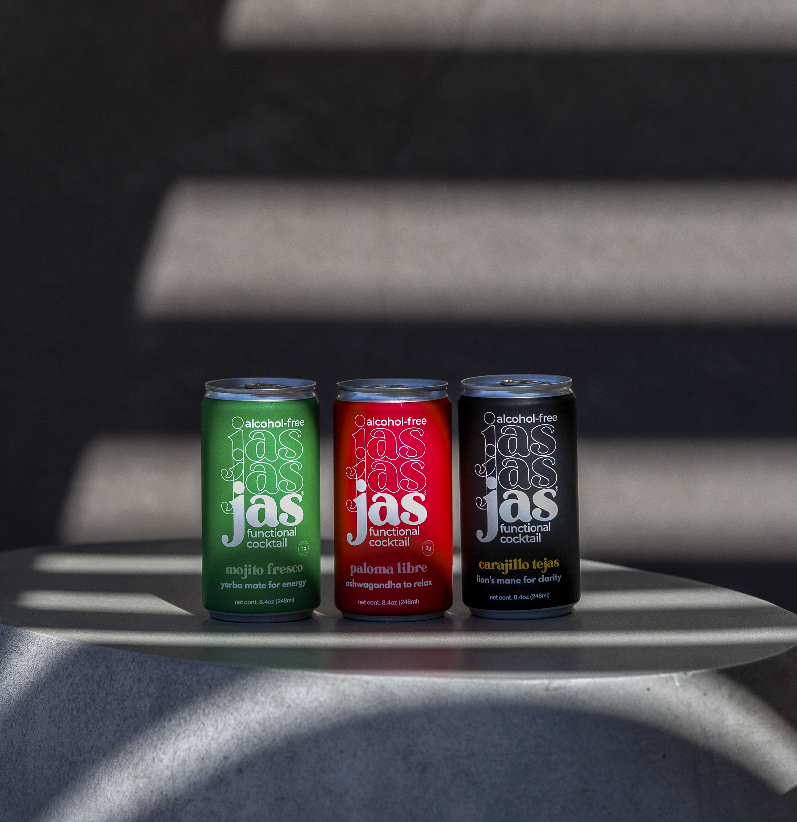 Three slim cans of Jas Non-alcoholic Cocktails sit on a round metallic table under dramatic diagonal shadows. The cans are green, red, and black, each with the brand name 'jas' in a stylized white script.