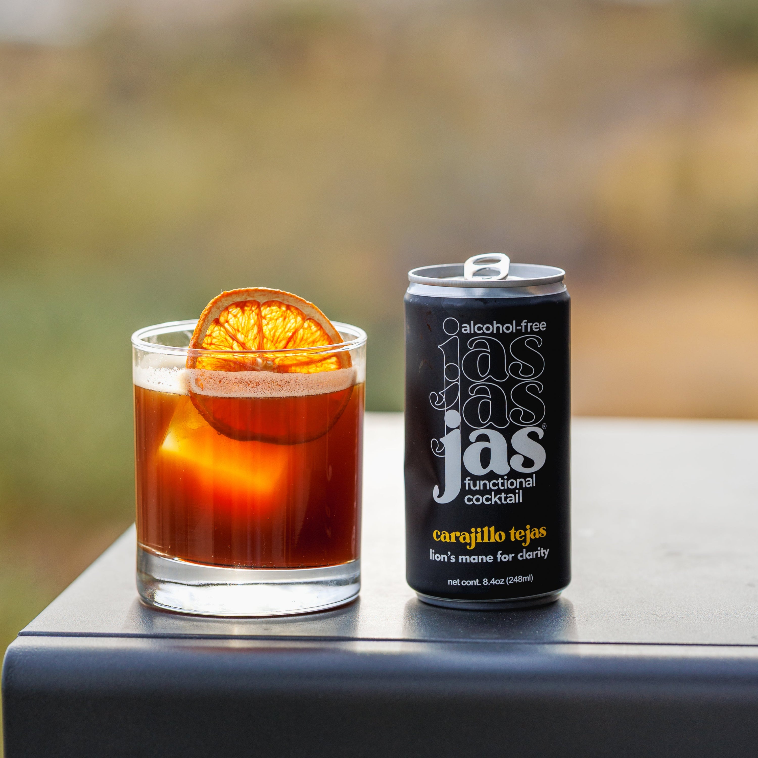 A black can of jas alcohol-free cocktail mix next to a glass of amber-colored cocktail with an orange slice garnish, set on a dark table with a blurred outdoor desert landscape background.