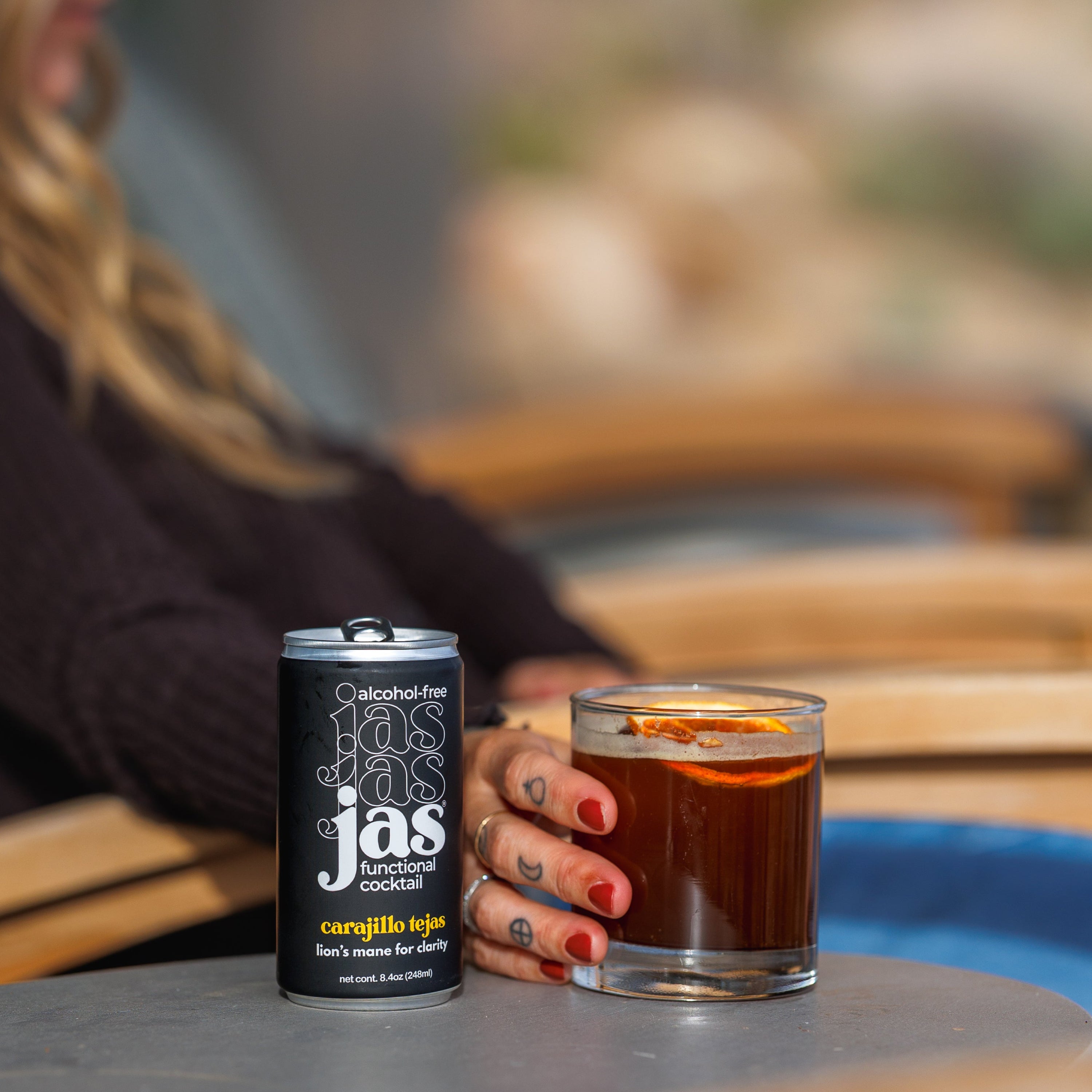 A person sitting outdoors by a table holds a glass of dark coffee next to a black can of jas coffee, with a blurred patio background.