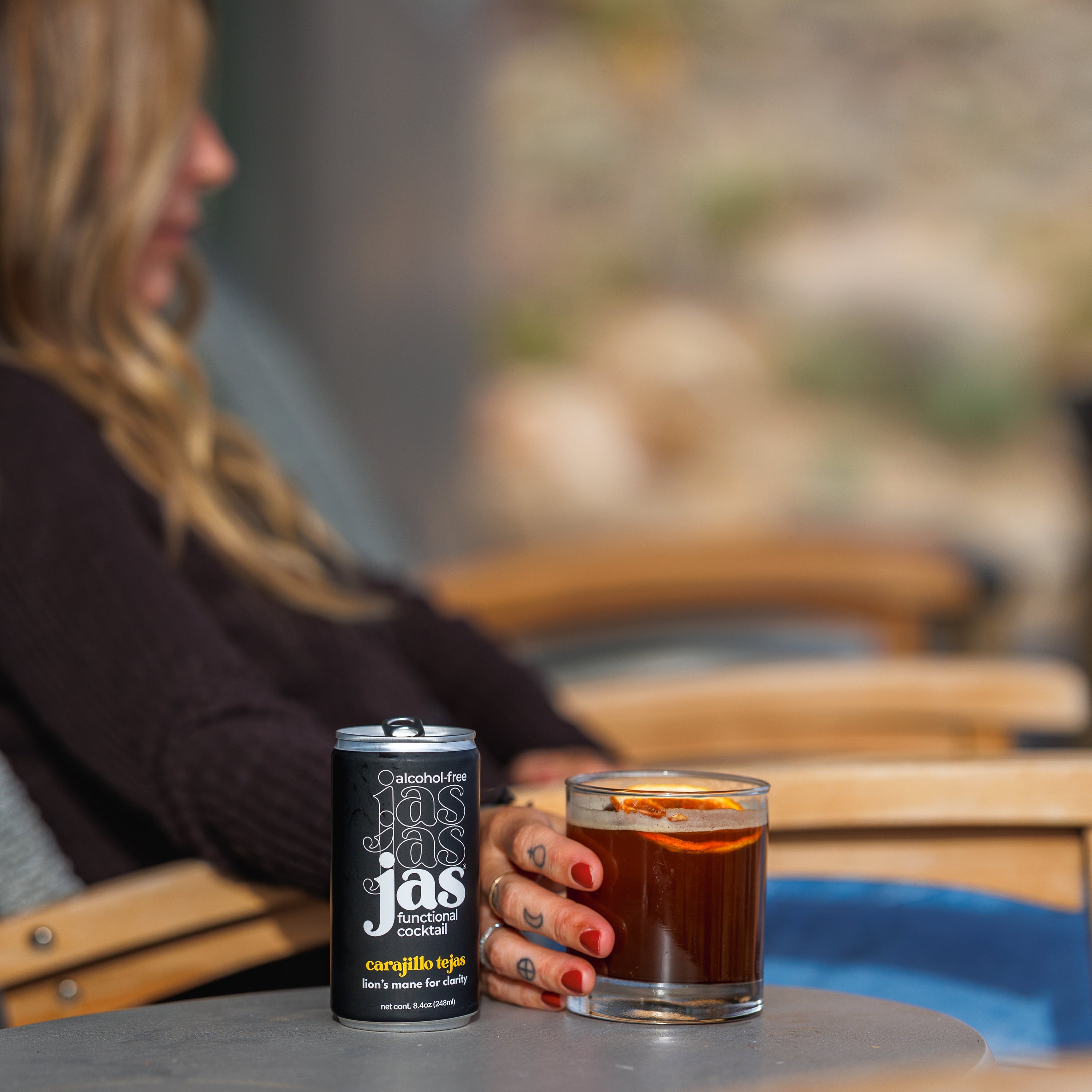 A person sitting outdoors by a table holds a glass of dark coffee next to a black can of jas coffee, with a blurred patio background.
